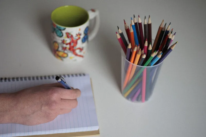 Een kinderhand schrijft in een schrift naast een beker kleurpotloden